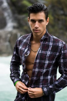 Handsome young man near mountain waterfall with open shirt, ripped athletic body
