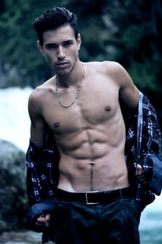 Handsome young man near mountain waterfall with open shirt, ripped athletic body