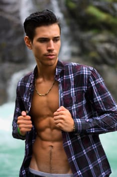 Handsome young man near mountain waterfall with open shirt, ripped athletic body