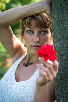 Attractive woman in a stylish white lacy top holding out a fragrant red rose symbolic of love as she leans against a tree in the garden
