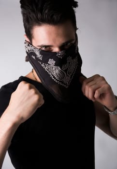Man with his face hidden behind a bandanna balling his fists aggressively and raising them threateningly as he stares balefully at the camera