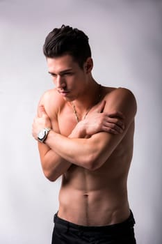 Handsome shirtless young man hugging himself while staring morosely at the floor as though comforting himself in his loneliness, on grey