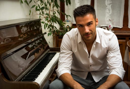 Young handsome male artist smiling while sitting next to his wooden classical upright piano, indoor portrait