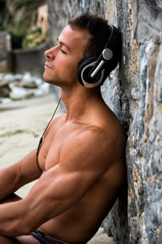 Solitary athletic young man leaning on a stone wall on the beach while listening to music through headphones