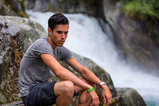 Handsome young man near mountain waterfall sitting on rocks, looking down