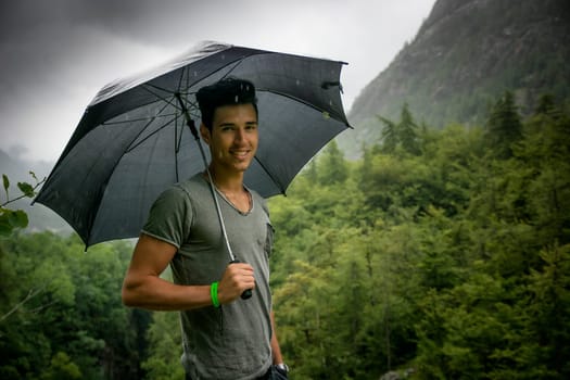 Smiling handsome young man holding an umbrella on a stormy day as he takes a walk along a trail in the mountains turning to smile at the camera