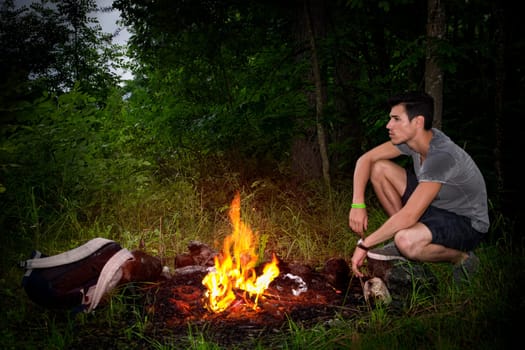 Young man camping in the mountains crouching alongside a burning campfire with his rucksack staring thoughtfully into the distance, profile view