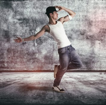 Hip young man in a tank top and hat doing a dance routine posing on one leg in a grunge concrete room or stage, profile full length view