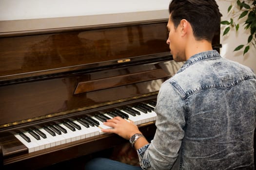 Young handsome male artist playing his wooden classical upright piano, indoor portrait