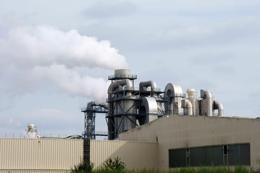 The photograph of a smouldering chimney as well as pipes and boilers of an industrial plant.