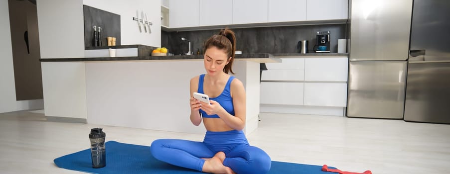 Portrait of sportswoman using sport app while doing workout, yoga exercises at home, sits on mat with water bottle, smiling and looking happy.