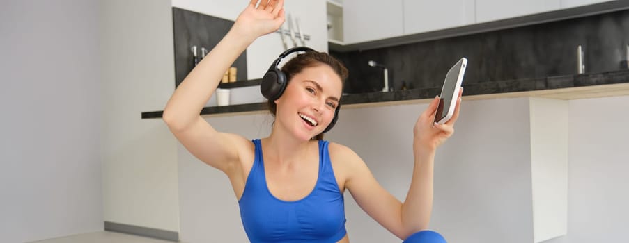 Happy fitness woman dancing, having fun during workout at home, wears headphones, holds smartphone, listens music during exercises.