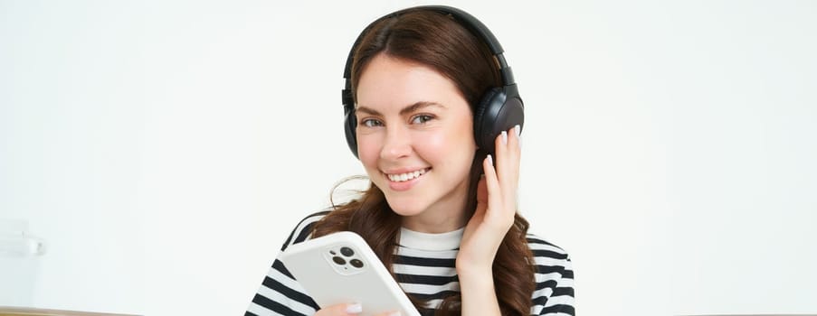 Portrait of happy young modern woman, female model in wireless headphones, smiling and looking at camera, holding smartphone, using mobile phone app, white background.