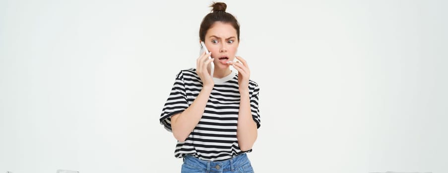 Image of woman with shocked face, looks confused while chats over the phone, listens to person on telephone, white background.