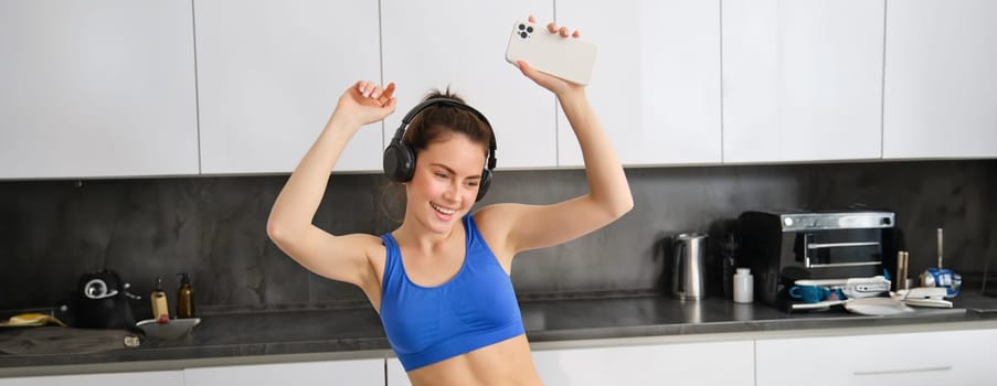 Portrait of carefree sportswoman, dancing in kitchen with smartphone, listening music in headphones, feeling energized and excited after workout.