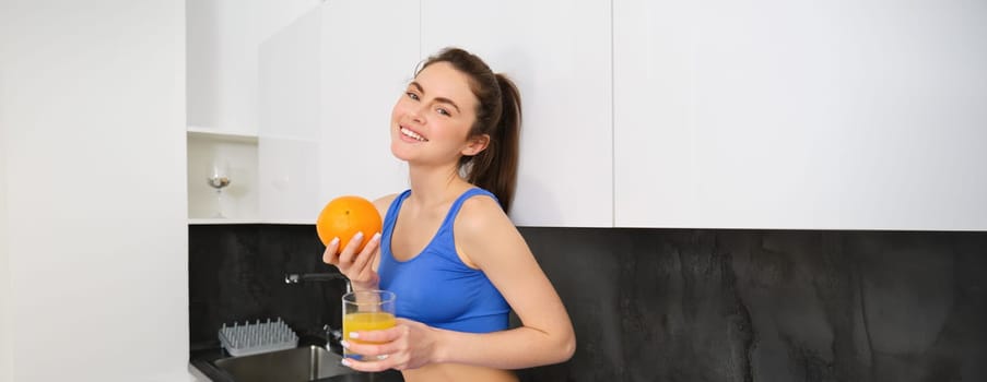 Indoor shot of woman after workout, standing in kitchen with fresh juice and an orange, drinking it.