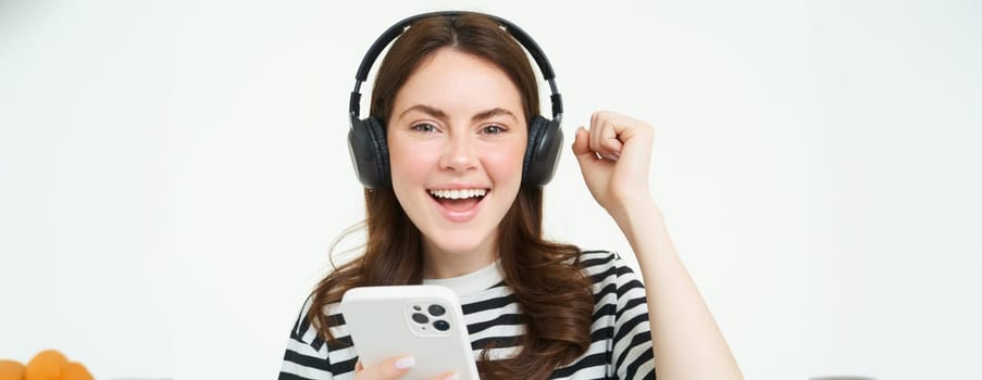 Enthusiastic girl in headphones, holding smartphone, smiling and cheering, celebrating, raising hand up, achieve goal, standing over white background.