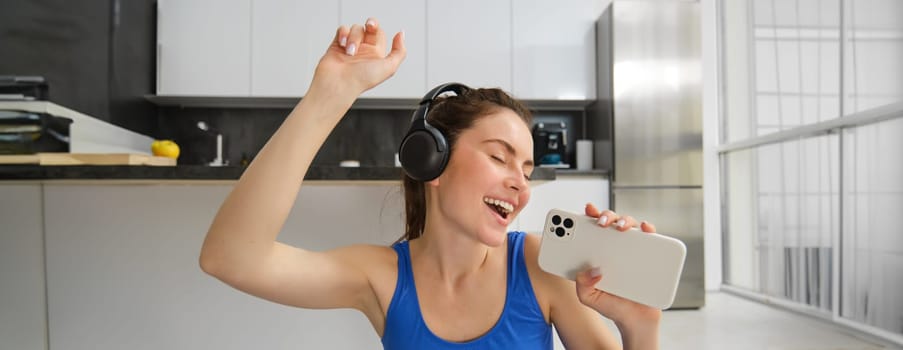 Happy young girl doing fitness, singing and having fun, listening music in headphones, holding smartphone, workout in living room.