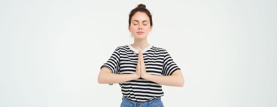 Portrait of young mindful woman meditating, holds hands clasped together, namaste gesture, practice yoga, standing over white background.
