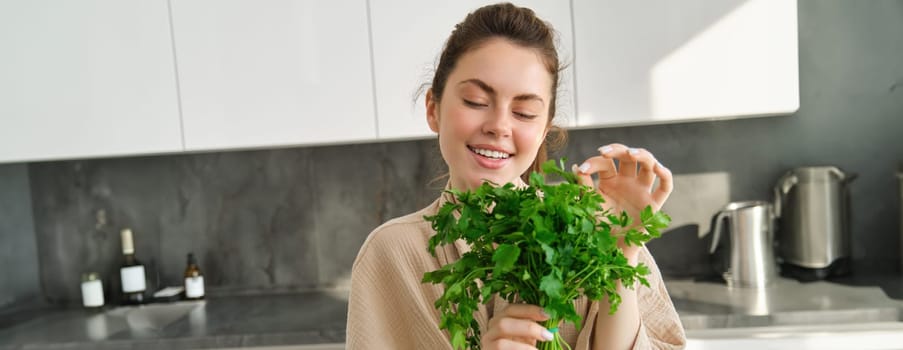 Portrait of beautiful woman cooking, holding fresh parsley, adding herbs while making salad, healthy meal in the kitchen.