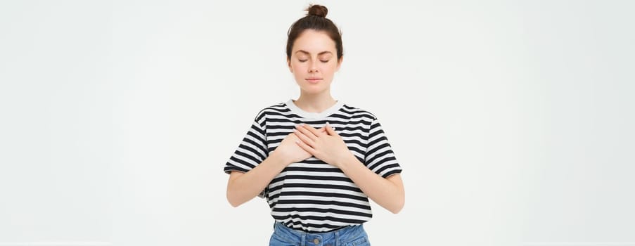 Portrait of young woman with calm, relaxed face, holds hands on heart with eyes closed, stands over white background.