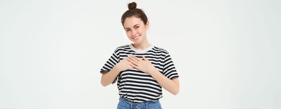 Young smiling woman, feels gratitude, thankful for something, holds hands on heart and looks tender at camera, standing over white background.