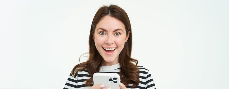 Portrait of excited brunette girl, reacting to amazing promo on smartphone, holding mobile phone and smiling, isolated on white background.