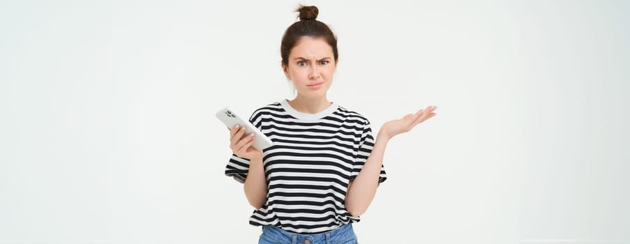 Confused woman shrugging shoulders, holds hands sideways, has phone in palm, clueless, frowning and looking puzzled, isolated over white background.
