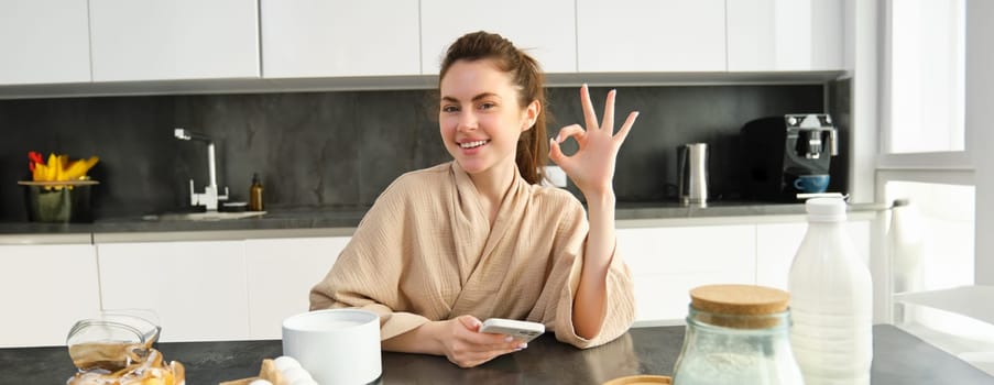 Attractive young cheerful girl baking at the kitchen, making dough, holding recipe book, having ideas.