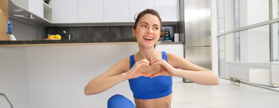Portrait of beautiful, fit young woman in leggins and sportsbra, sits in lotus pose on yoga mat at home, shows heart sign and smiles at camera, workout, fitness instructor online video training.