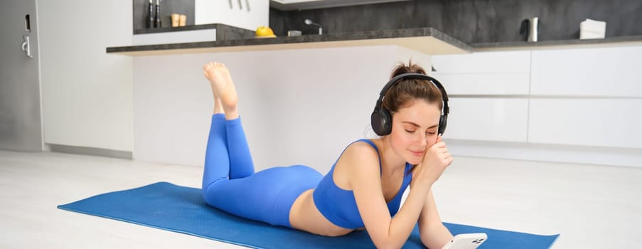 Portrait of brunette fitness girl, laying on yoga mat at home, watching workout tutorials, videos on smartphone, wearing sportswear and wireless headphones.