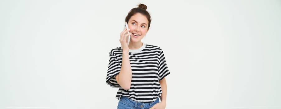 Happy young woman talks on mobile phone, chats on telephone, uses smartphone, stands over white background. Copy space