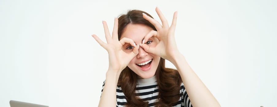 Portrait of happy, beautiful young woman, makes glasses with fingers around eyes and laughing, having fun, enjoying event, standing over white background.