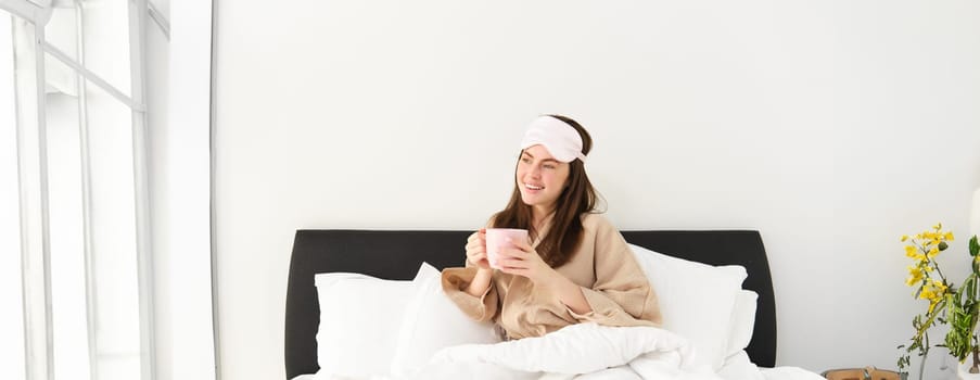Portrait of beautiful woman in sleeping bag and pyjamas, drinking coffee in bed, enjoying her bright, relaxing morning.