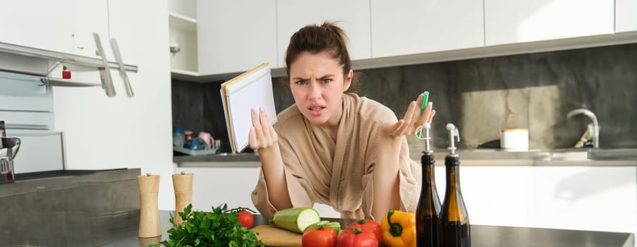 Portrait of woman cant cook, looking confused while making meal, holding recipe book, checking grocery list and staring frustrated at camera, standing near vegetables in the kitchen.