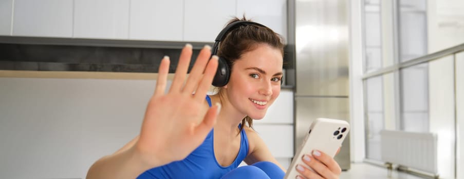 Happy fitness instructor at home, does workout with headphones and smartphone, listens to music and doing training exercises.