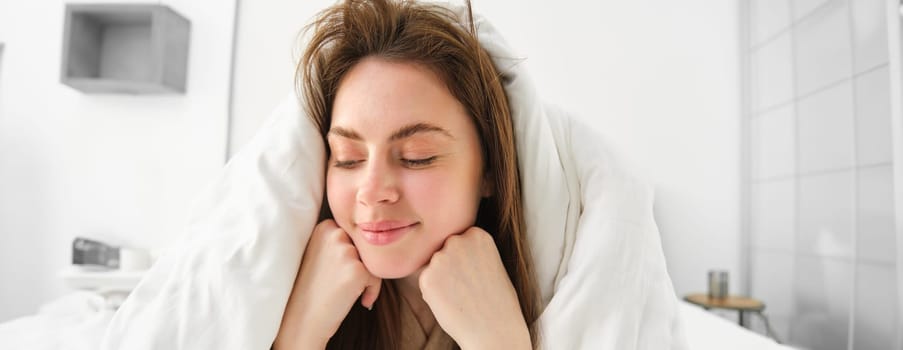 Cheerful woman feeling comfortable in bed, lying in bedroom covered in white sheets, smiling pleased, has messy hair in morning, waking up and looking happy.