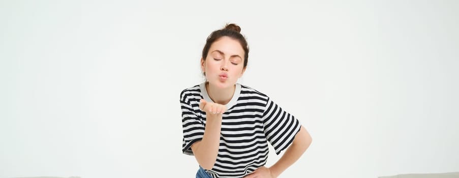 Portrait of beautiful brunette woman sends air kiss at camera, holds palm near lips, isolated against white background.