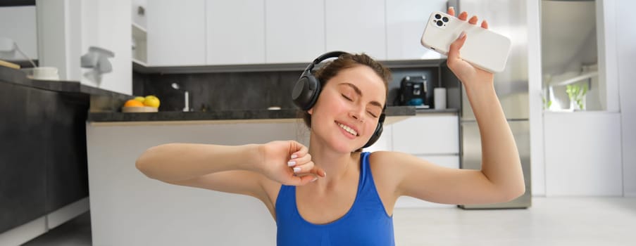 Happiness, sports and wellbeing. Young woman dancing in headphones, holding smartphone, doing workout at home, fitness training inside her house.