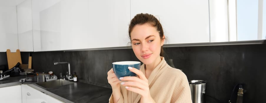 Portrait of happy young woman starts her morning with mug of coffee, drinking tea from cup, standing in the kitchen, smiling cheerfully.