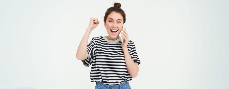 Enthusiastic young woman laughing, answers phone call and celebrating, receiving good news over the telephone, isolated over white background.