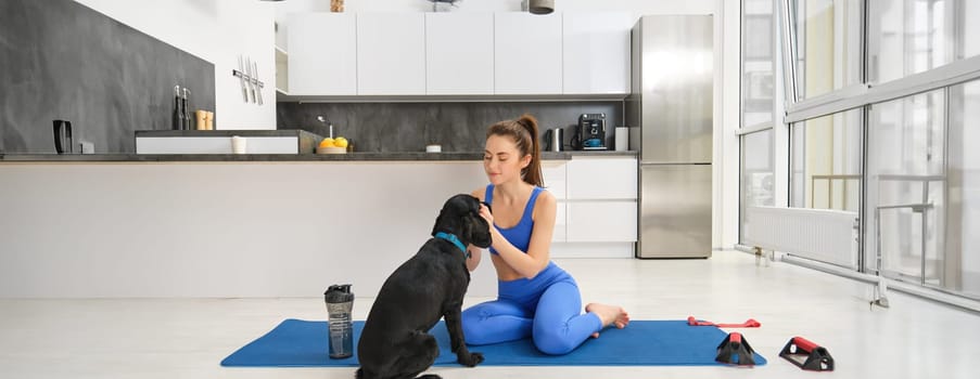 Image of young woman does workout from home, sits on yoga mat in living room and plays with her black dog, puppy distracts girl from doing exercises.