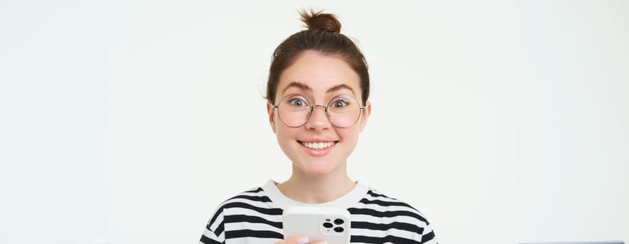 Portrait of beautiful young woman in eyewear, holding smartphone, using mobile phone, online shopping in application, standing over white background.
