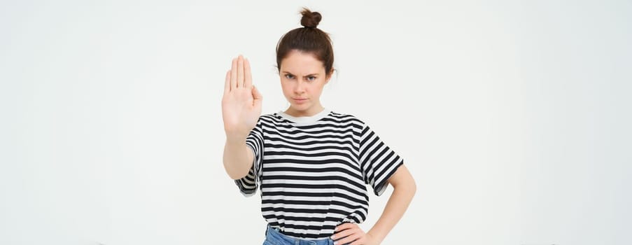 Image of woman frowning, showing one palm, stop gesture, disapprove and reject something, makes prohibit gesture, standing over white background.