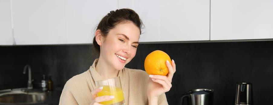 Close up portrait of stylish modern woman, drinking fresh juice from glass in kitchen, holding an orange, laughing and smiling, standing at home in bathrobe.