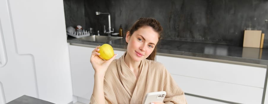Close up portrait of smiling brunette woman in bathrobe, sits in kitchen at home, uses mobile phone and holds an apple, orders fresh fruits on smartphone app, buys groceries online, looks up recipe.