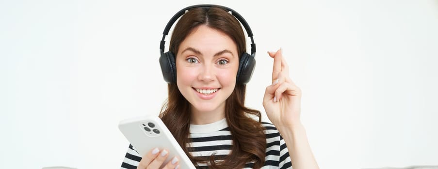 Young woman holding fingers crossed, making wish, hoping for something, holding smartphone, wearing wireless headphones, white background.