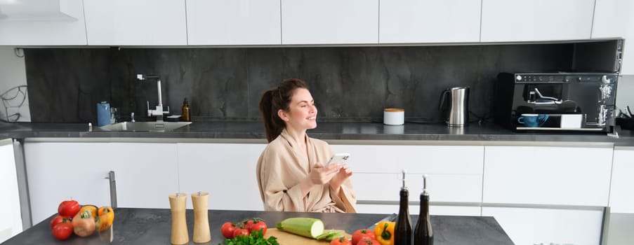 Portrait of woman in bathrobe sitting in kitchen with smartphone, cooking dinner, watching recipe on social media, video tutorial how to prepare meal.