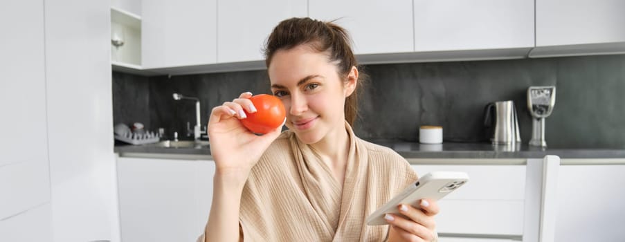 Close up portrait of beautiful smiling woman, holding fresh tomato, sitting in kitchen with smartphone, orders vegetables online, using application to buy groceries, using mobile phone.