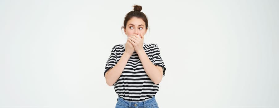 Image of shocked woman, covers her mouth, looks surprised, stands over white background. Copy space